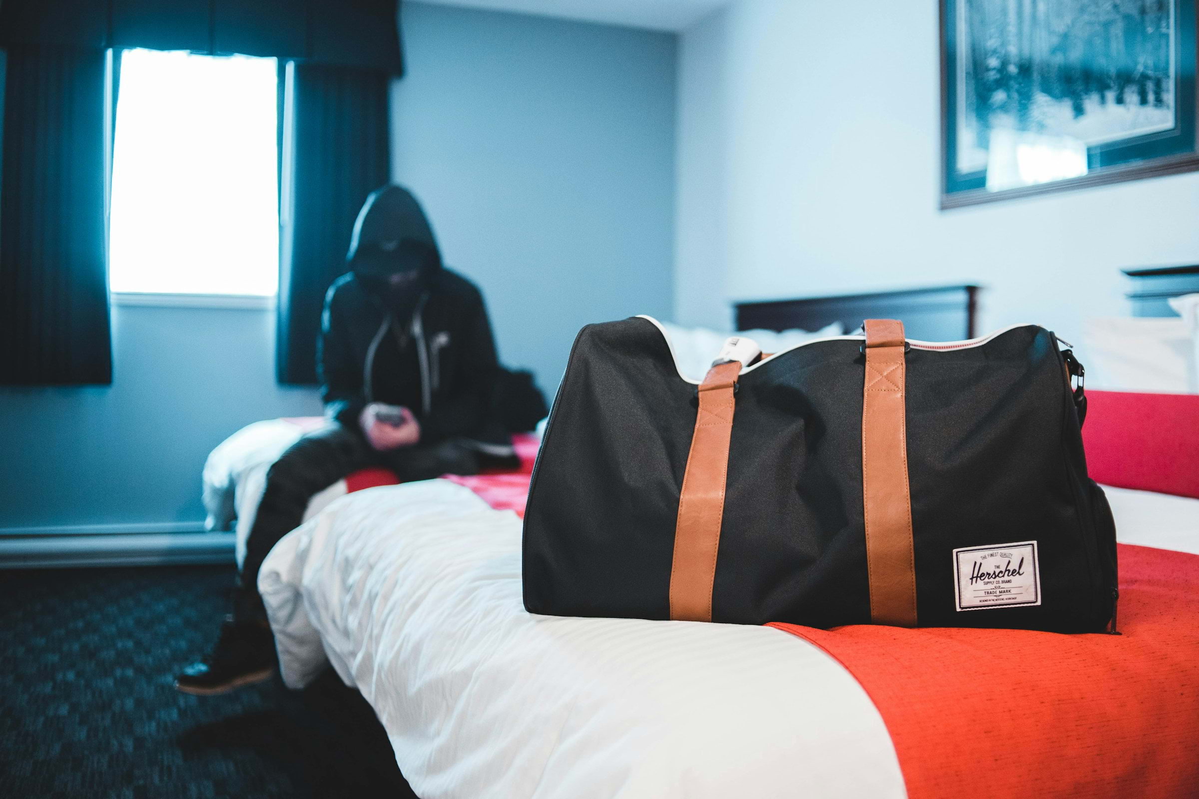 Man in black hooded jacket sitting on a hostel bed near a black-and-brown tote bag