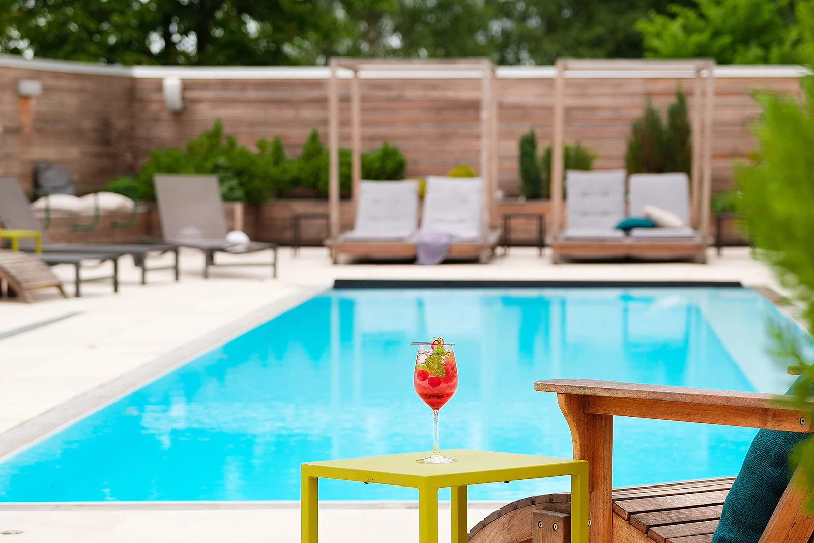 A red drink on a green table by a pool surrounded by sunbeds