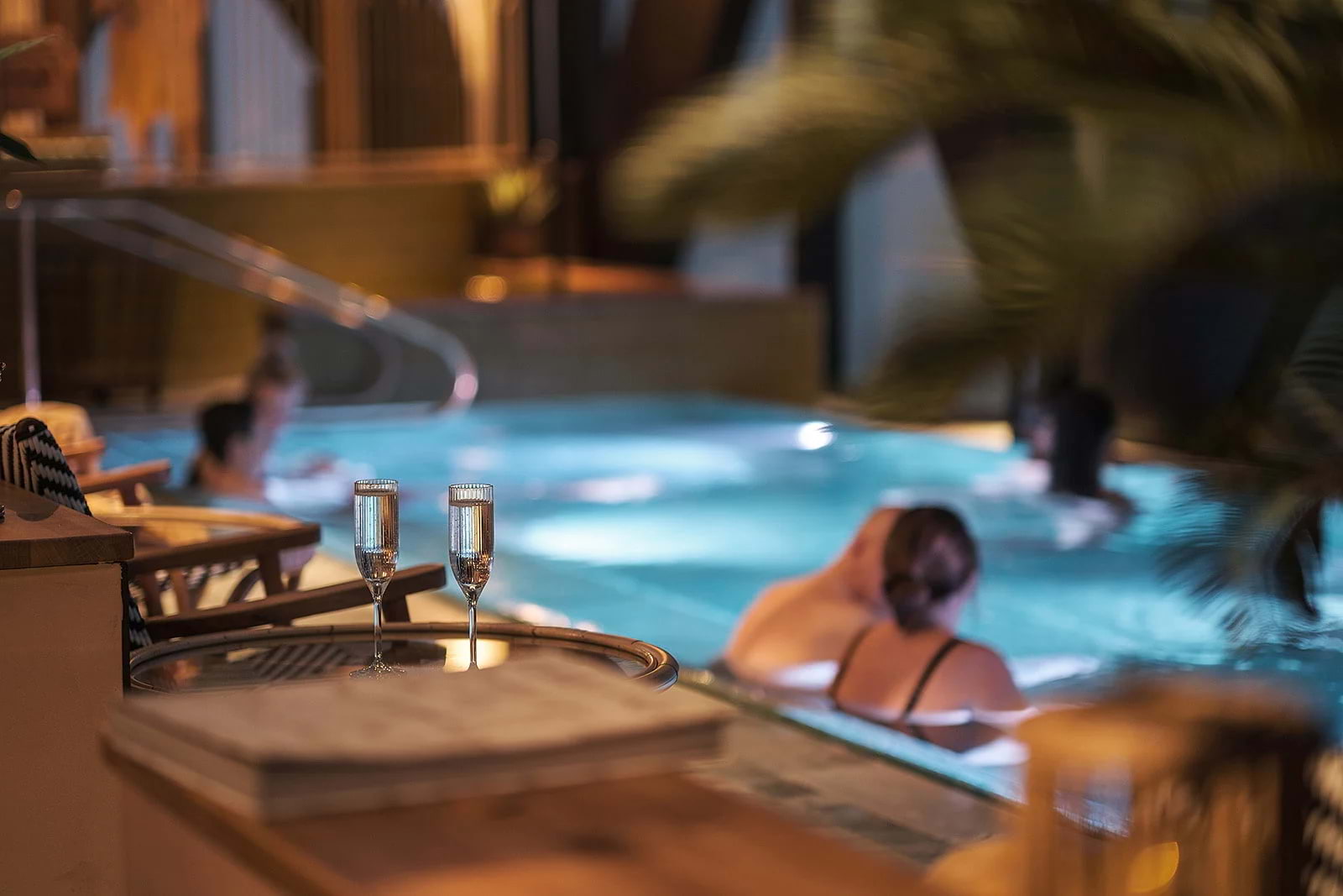 A pair of champagne galsses set out by a pool, with a couple in the water in the background