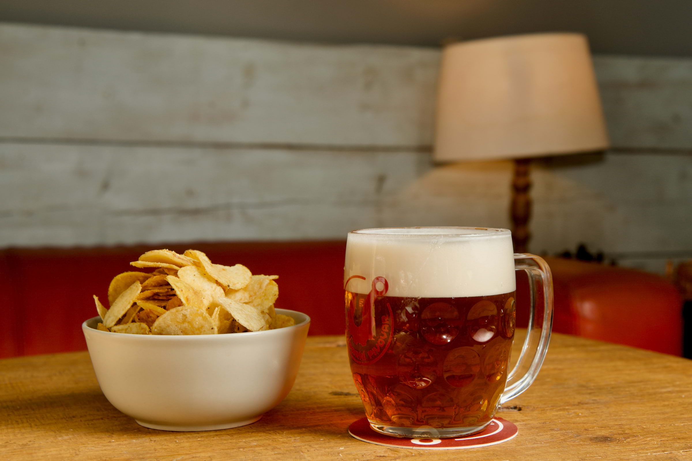 A bowl of crisps next to a pint of beer on a wooden table with a white lamp in the background