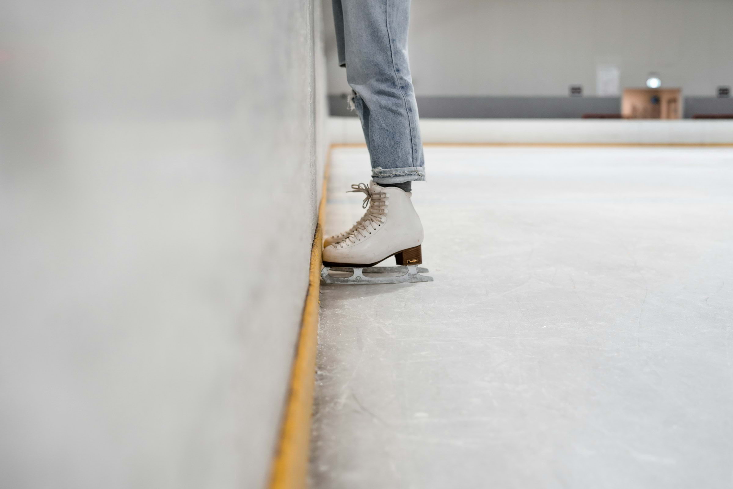 A person wearing grey jeans and white ice skates at the edge of an indoor ice-skating rink