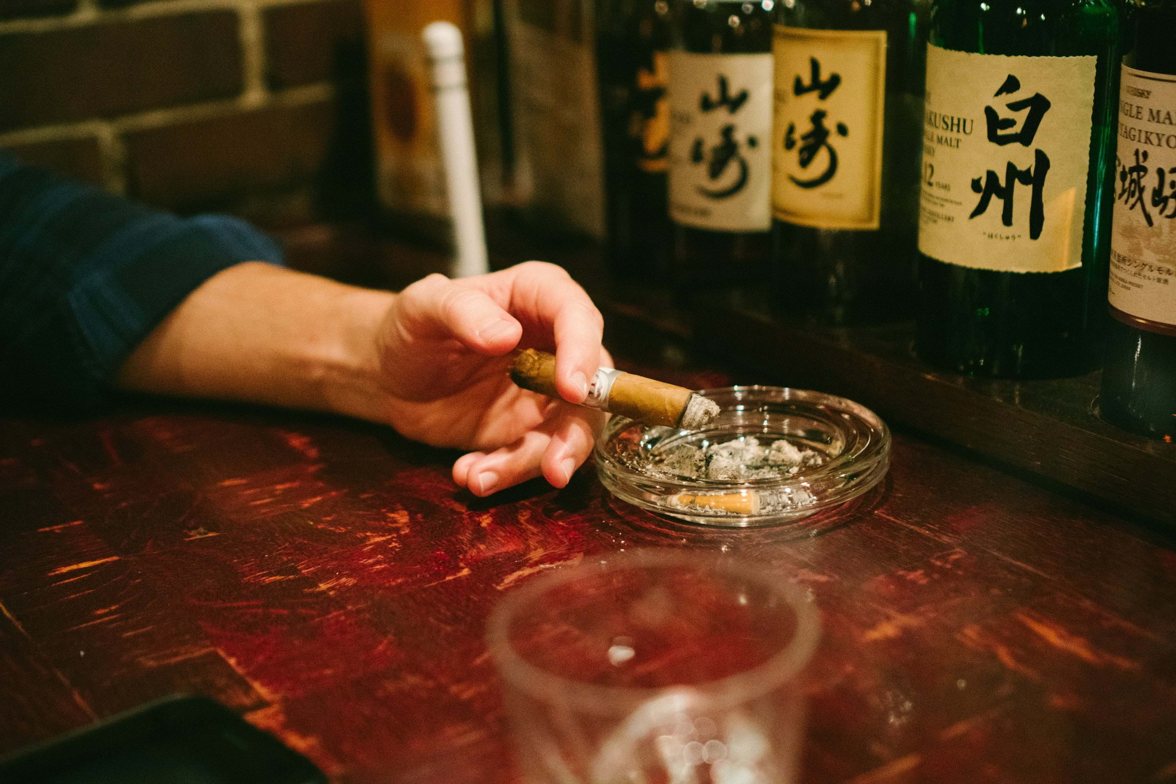 A man's hand holding a lit cigar over an ashtray, with Japanese whiskies in the background