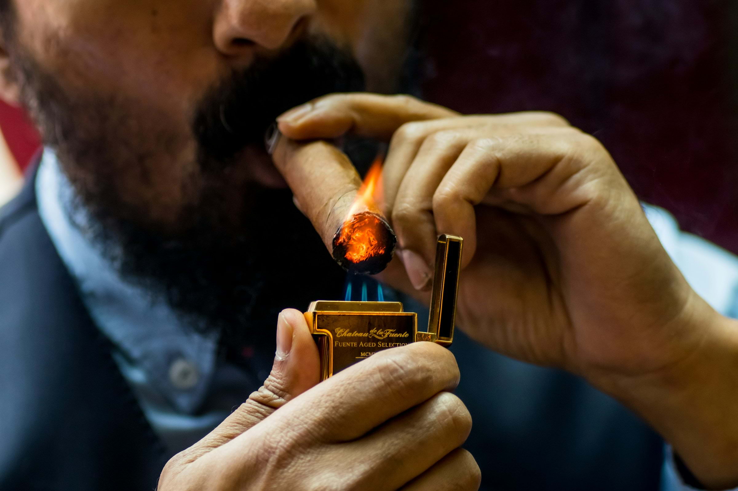 A close-up of a bearded man lighting a cigar with a golden lighter