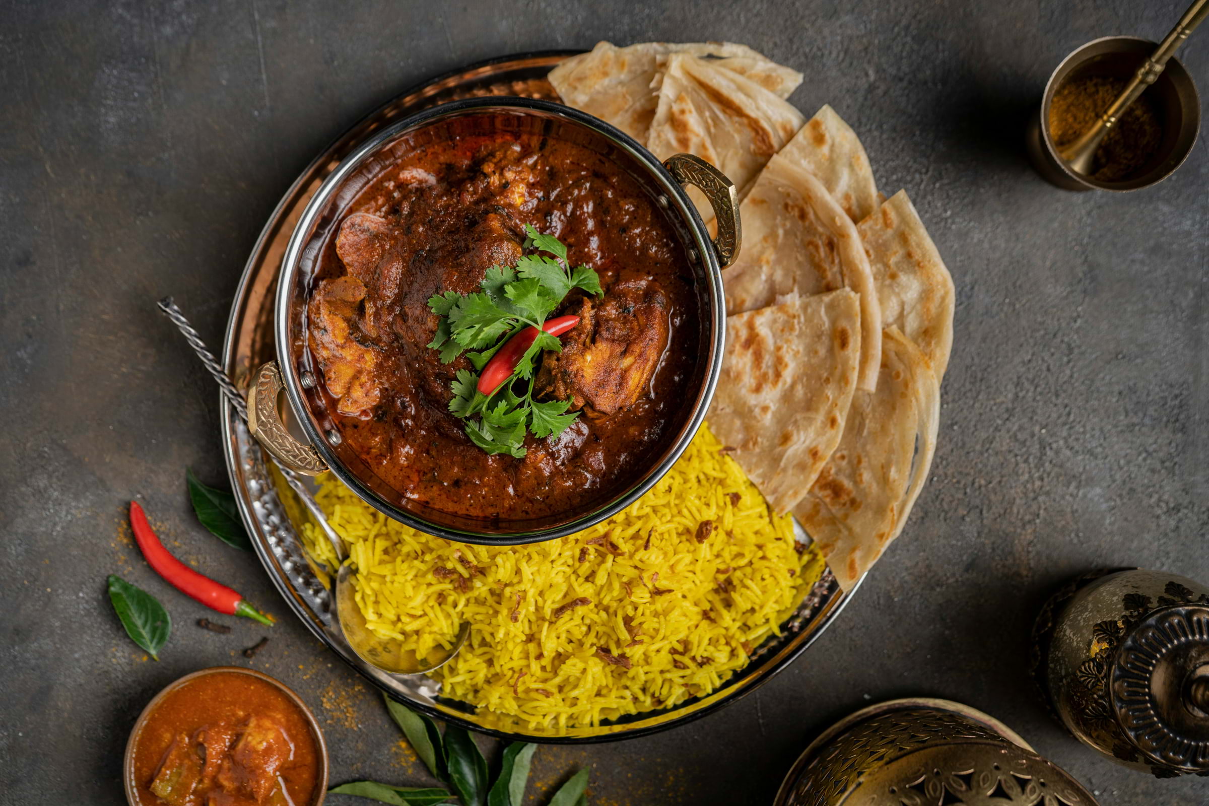 A bird's eye view of a dark brown meaty curry next to paratha and pilau rice.