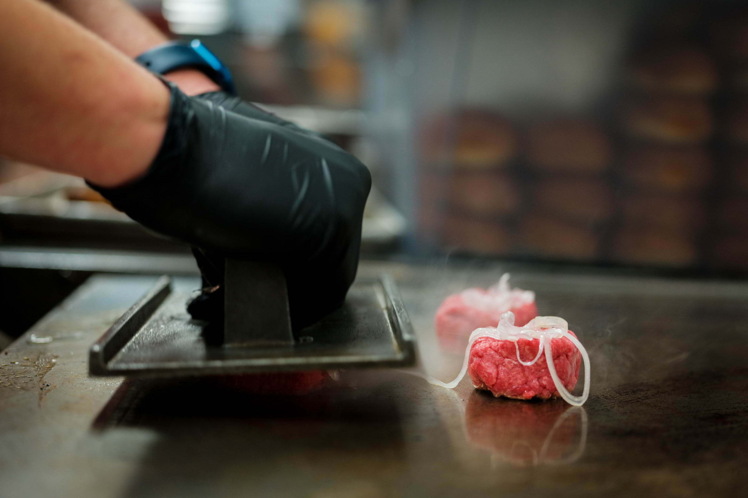 A cook in black gloves smashing a burger patty on a grill
