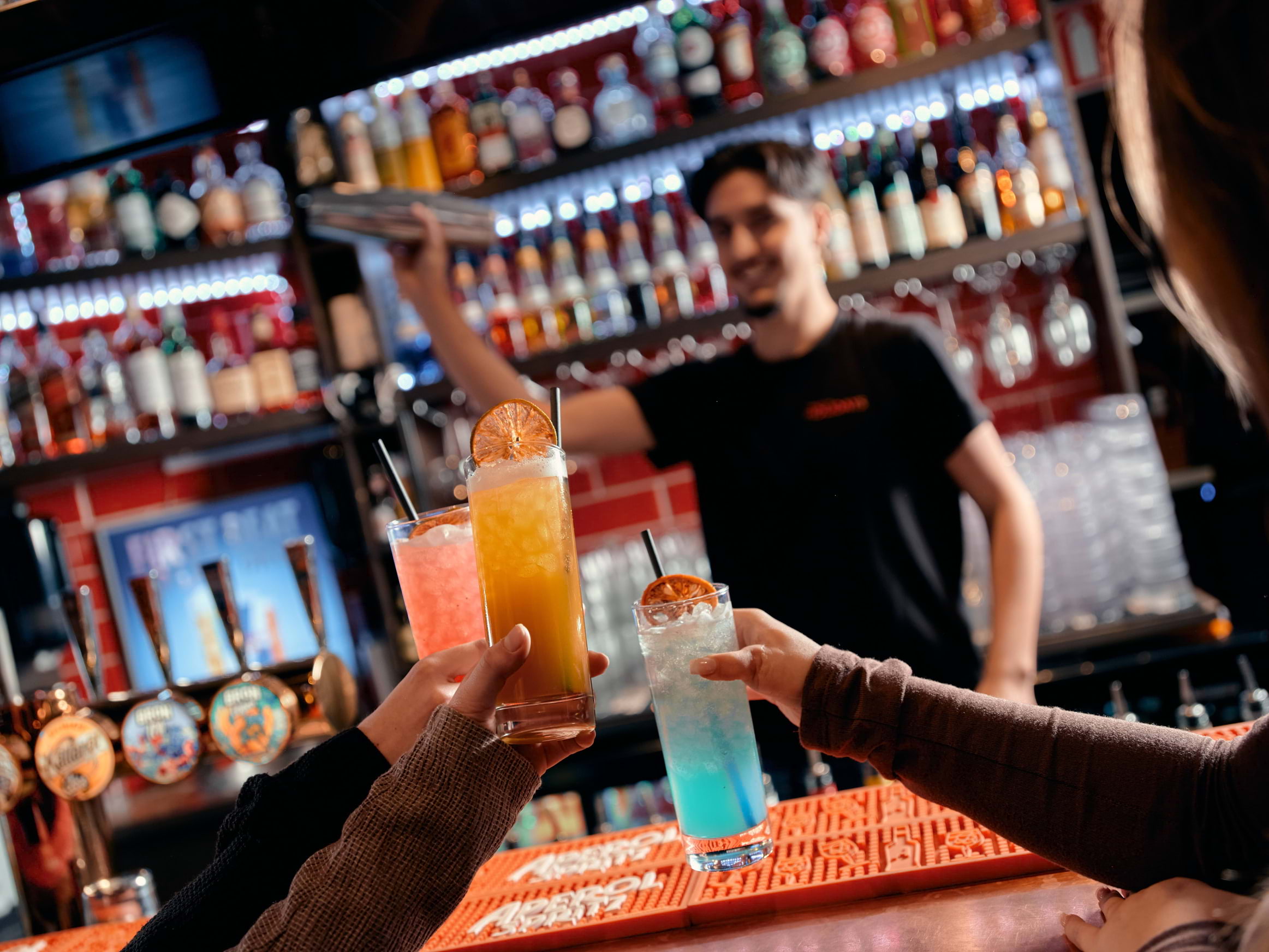 Three people toasting with different-coloured cocktails in front of a bartender