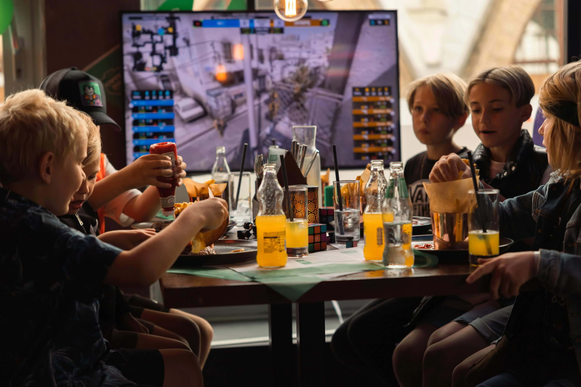 A group of kids eating at a restaurant table with a gaming screen in the background