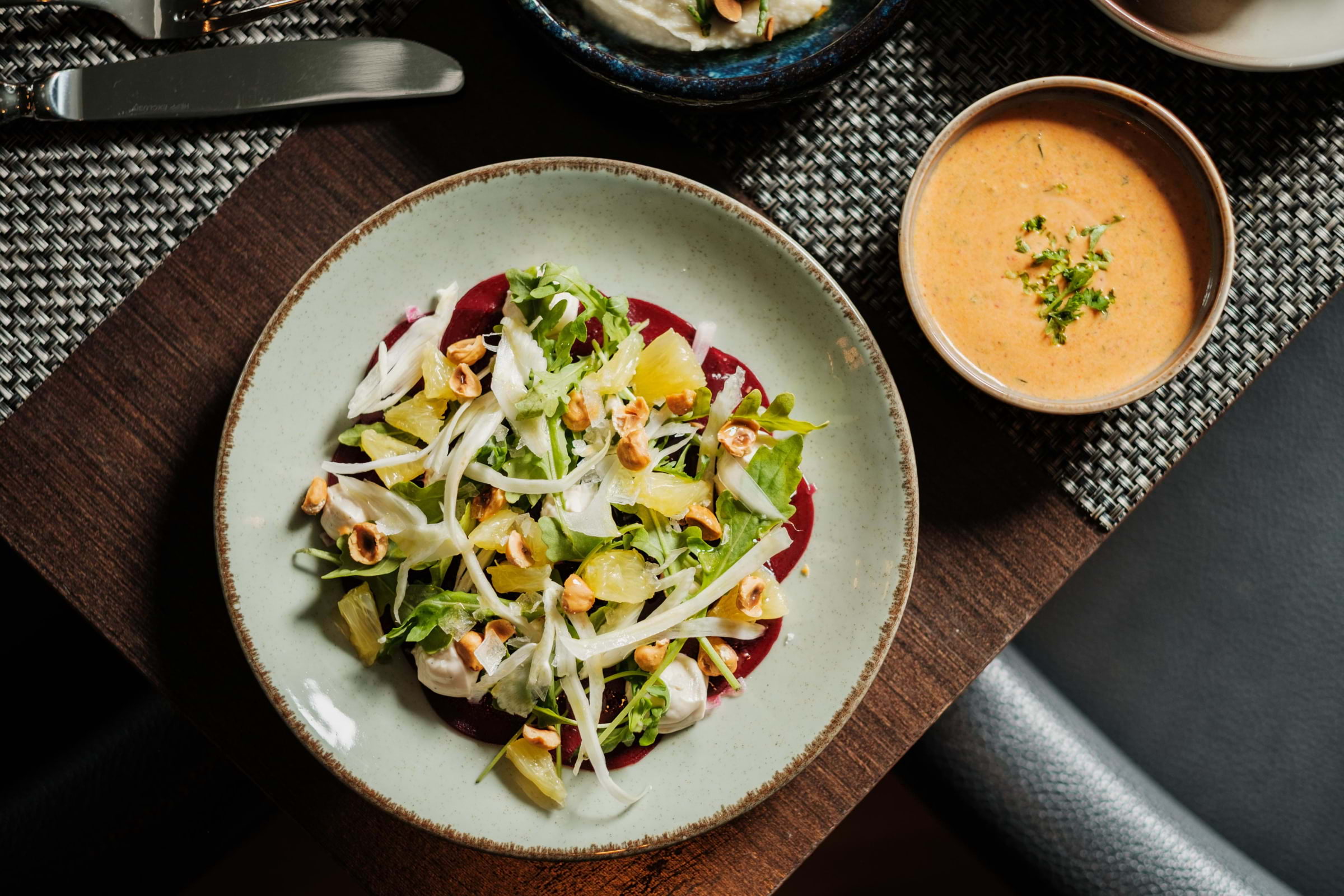A salad on a bespoke ceramic plate and a small bowl of orange soup on a restaurant table