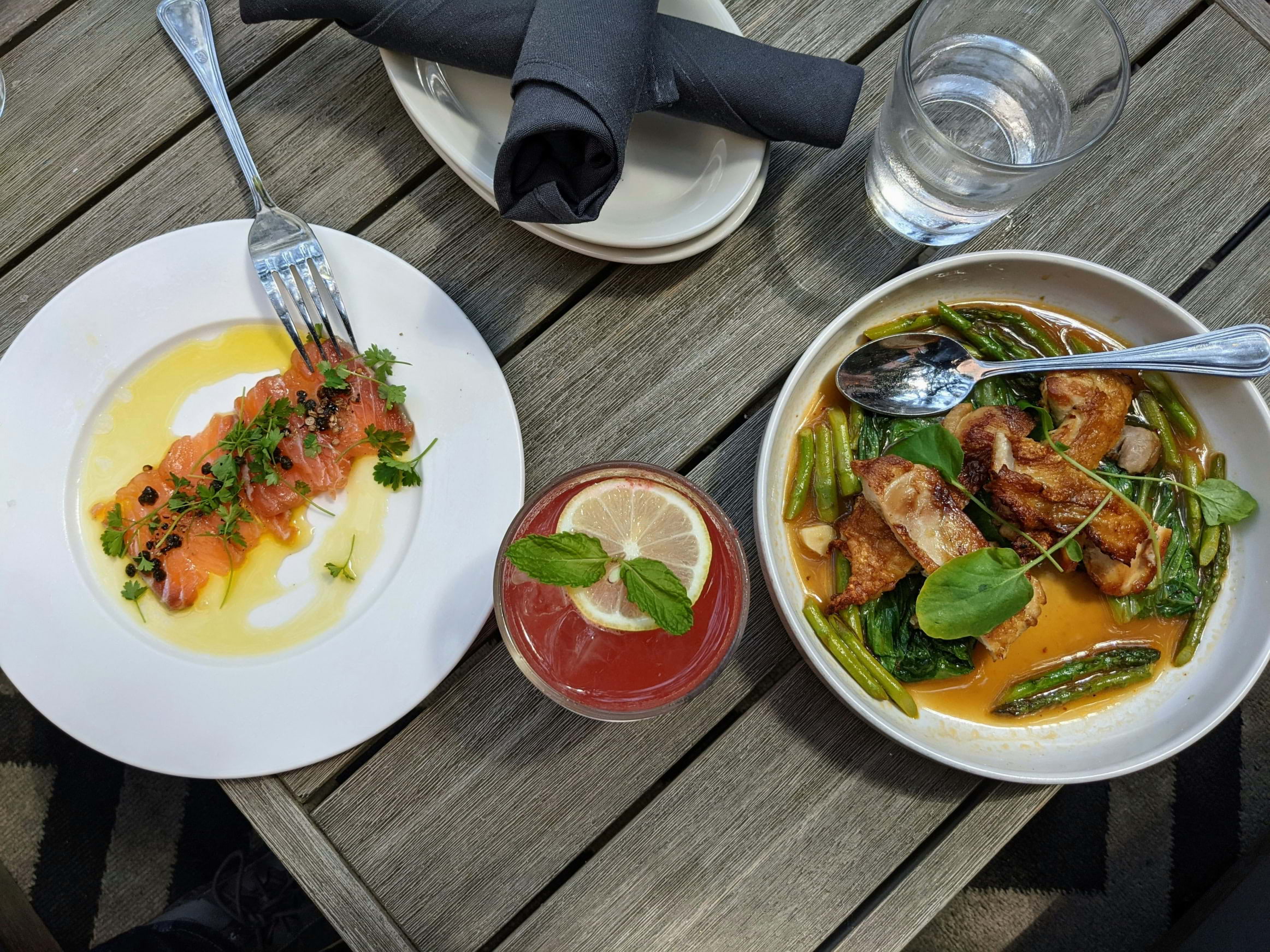 Two plates of food on a dark wooden table with a pink cocktail