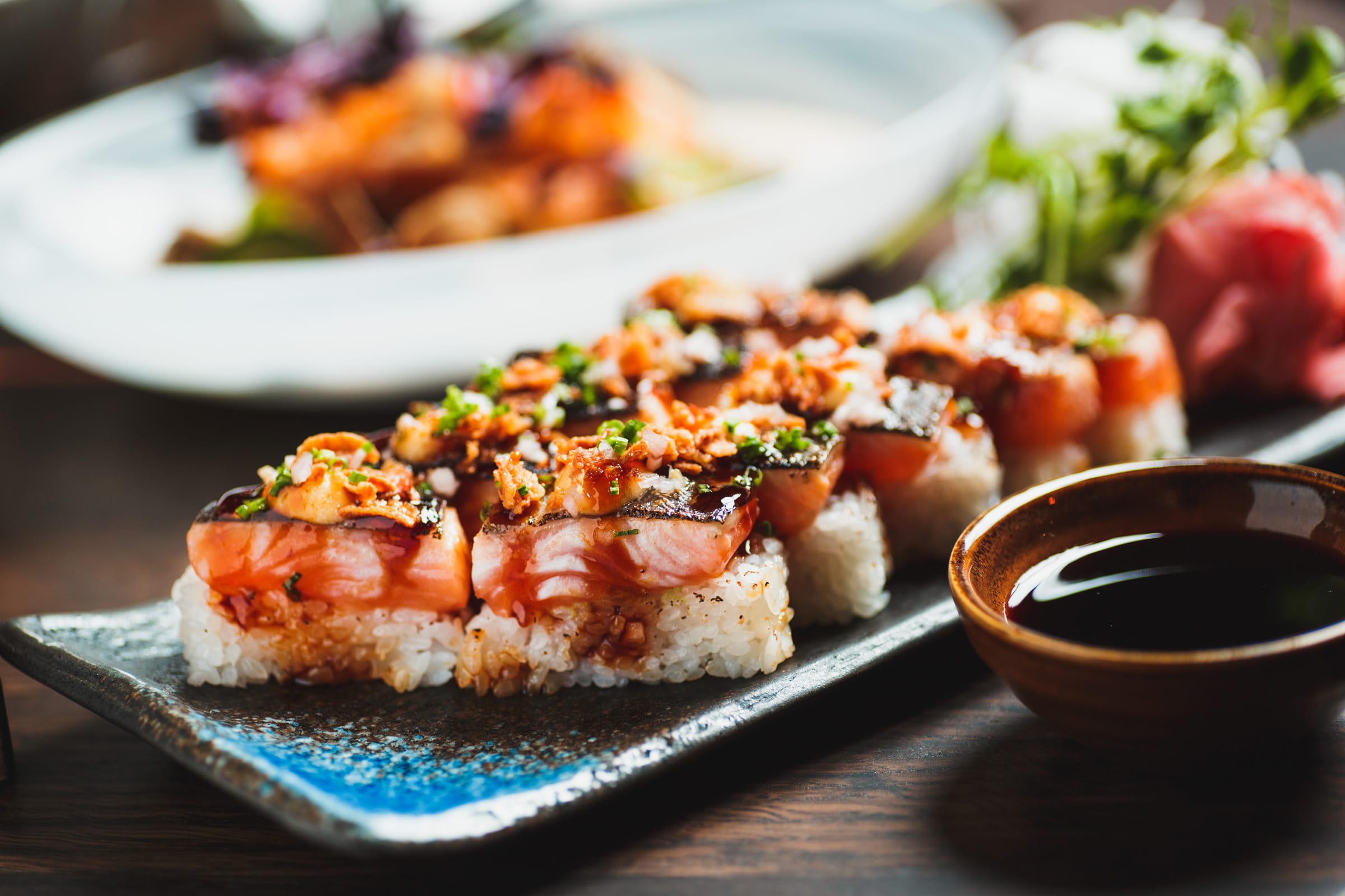 En tallrik med sushi och sojasås i en skål på sidan och en suddig tallrik med färgglad mat i bakgrunden